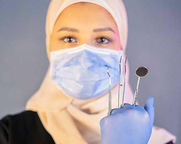 "A professional female dentist, Dr. Sarah Al-Dandal, in a dental clinic setting, wearing a hijab and dental uniform with gloves on, standing next to dental equipment."